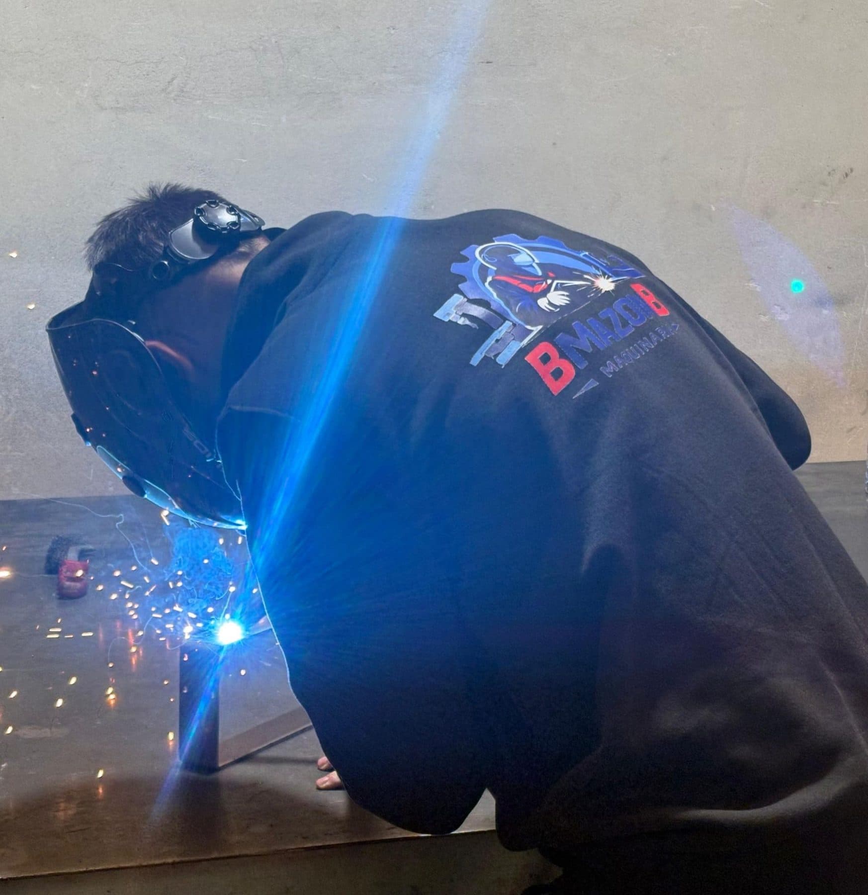 Borja soldando con MIG entre chispas, con el logo de BMAZONB en la sudadera